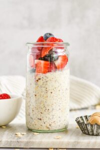 Overhead shot of three glass mason jars filled with creamy overnight oats layered with fresh strawberries, blueberries, and raspberries, topped with a drizzle of honey and fresh mint leaves on a light linen surface