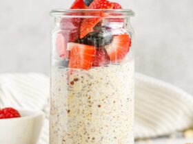 Overhead shot of three glass mason jars filled with creamy overnight oats layered with fresh strawberries, blueberries, and raspberries, topped with a drizzle of honey and fresh mint leaves on a light linen surface