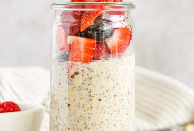 Overhead shot of three glass mason jars filled with creamy overnight oats layered with fresh strawberries, blueberries, and raspberries, topped with a drizzle of honey and fresh mint leaves on a light linen surface