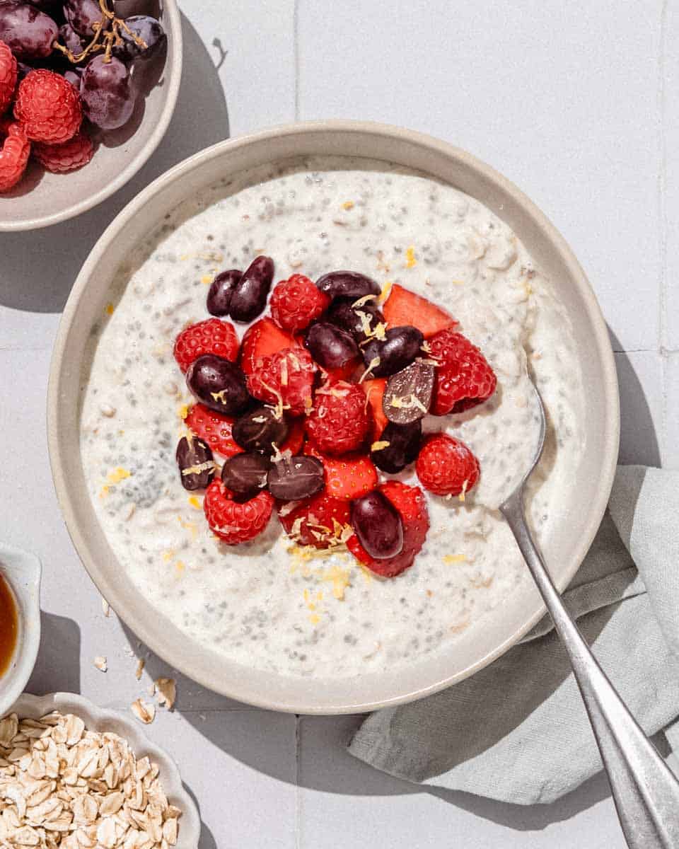 Flat lay ingredients shot showing rolled oats in a small bowl, glass of milk, container of Greek yogurt, fresh mixed berries in a colander, maple syrup, vanilla extract, and chia seeds arranged on a white marble surface