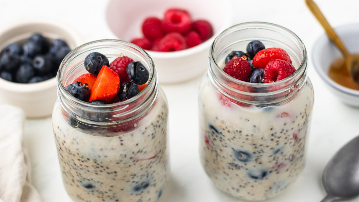 Close-up shot of a single mason jar of overnight oats with summer berries, showing distinct layers of creamy oats and vibrant mixed berries, topped with a golden honey drizzle and a sprig of fresh mint, natural morning light