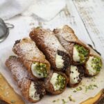 Golden crispy cannoli shells filled with rich white ricotta cream, garnished with chopped pistachios and chocolate chips, arranged on an elegant marble serving plate