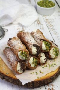 Golden crispy cannoli shells filled with rich white ricotta cream, garnished with chopped pistachios and chocolate chips, arranged on an elegant marble serving plate