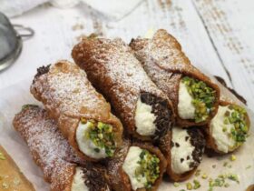 Golden crispy cannoli shells filled with rich white ricotta cream, garnished with chopped pistachios and chocolate chips, arranged on an elegant marble serving plate