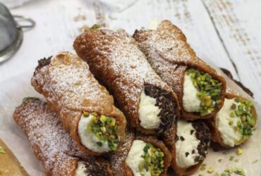 Golden crispy cannoli shells filled with rich white ricotta cream, garnished with chopped pistachios and chocolate chips, arranged on an elegant marble serving plate
