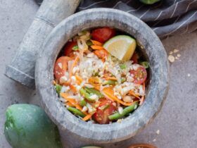 Beautifully plated Thai-style green papaya salad som tam in a white bowl with shredded papaya, tomatoes, green beans, roasted peanuts, and lime wedges on a wooden table