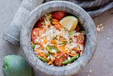 Beautifully plated Thai-style green papaya salad som tam in a white bowl with shredded papaya, tomatoes, green beans, roasted peanuts, and lime wedges on a wooden table
