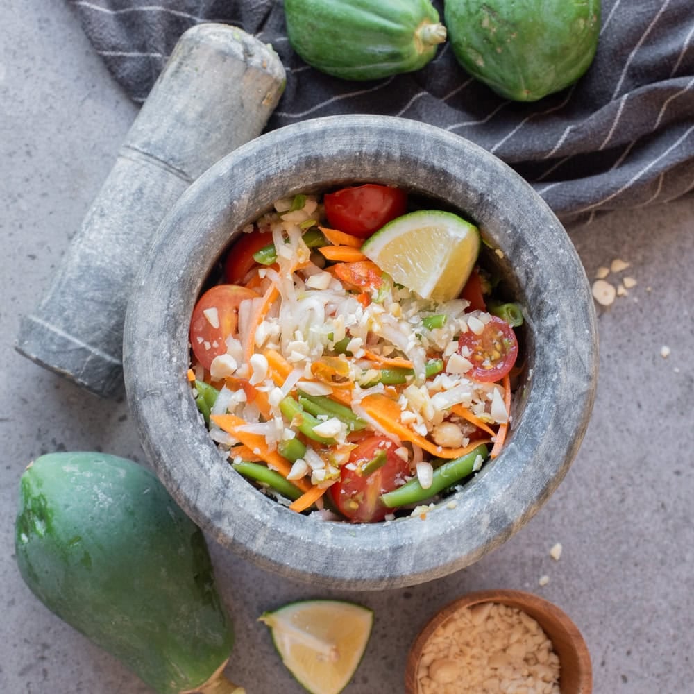 Beautifully plated Thai-style green papaya salad som tam in a white bowl with shredded papaya, tomatoes, green beans, roasted peanuts, and lime wedges on a wooden table