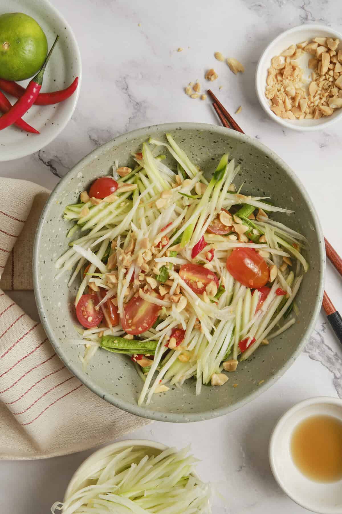 Overhead flat lay of Thai papaya salad ingredients including shredded green papaya, bird's eye chillies, limes, fish sauce, palm sugar, cherry tomatoes, green beans, and roasted peanuts on a marble surface