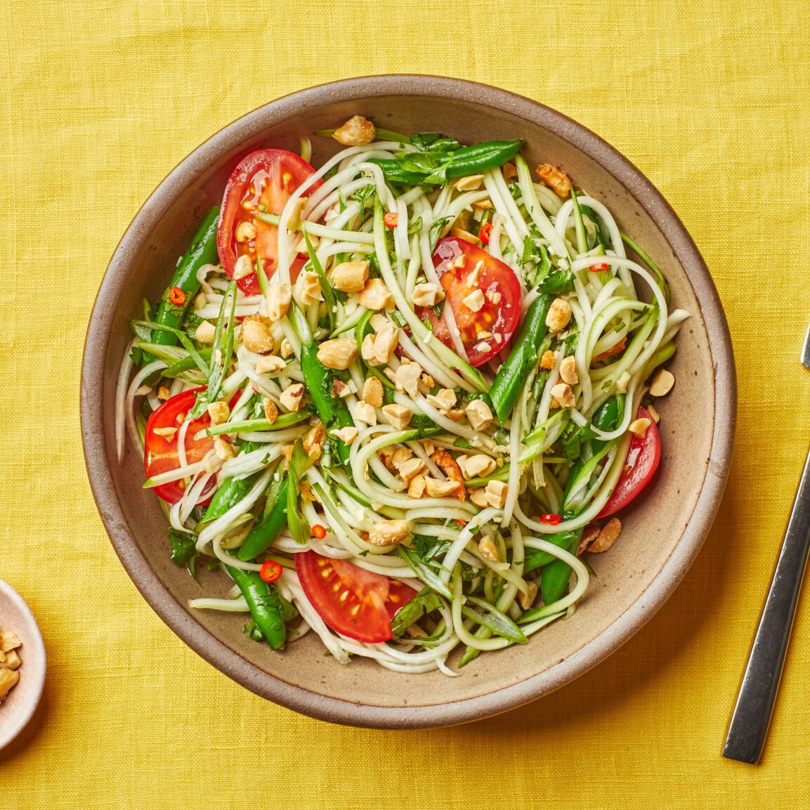 Close-up shot of Thai papaya salad som tam in a rustic bowl showing shredded green papaya strands, bright red cherry tomatoes, crushed peanuts, and a lime wedge with fresh herbs scattered over the top