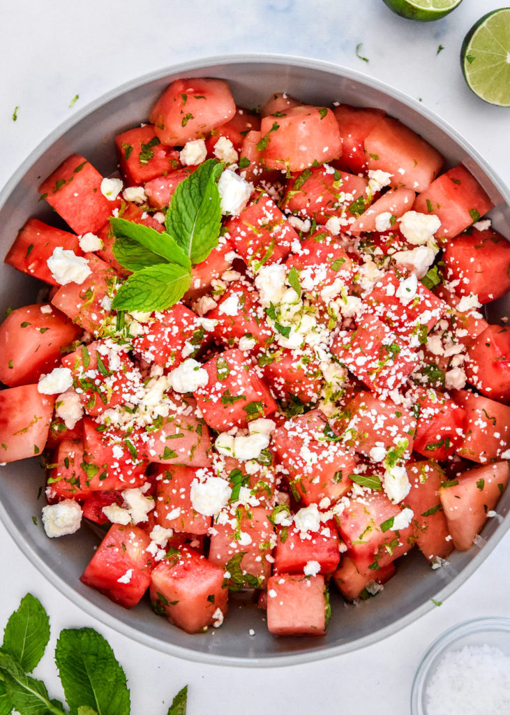 Step-by-step preparation shot showing cubed watermelon, hand-crumbled feta cheese, fresh mint leaves, and lime wedges arranged on a marble cutting board
