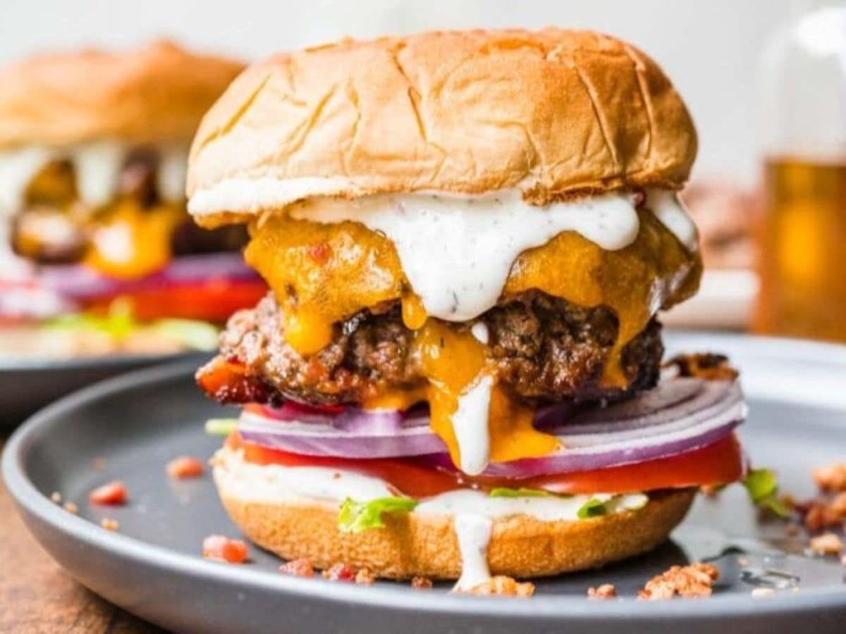 Bacon Ranch Burgers stacked high on a brioche bun with melted cheddar, crispy bacon, fresh lettuce, tomato, and a drizzle of ranch dressing on a rustic wooden board