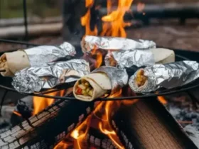 Campfire breakfast burritos wrapped in foil and resting on a campfire grate over glowing orange embers, one unwrapped burrito showing layers of scrambled eggs, sausage, and melted cheese