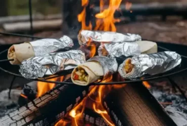 Campfire breakfast burritos wrapped in foil and resting on a campfire grate over glowing orange embers, one unwrapped burrito showing layers of scrambled eggs, sausage, and melted cheese