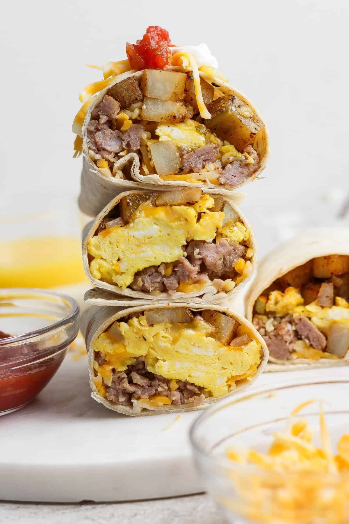 Flat lay of campfire breakfast burrito ingredients on a wooden cutting board: sausage, cracked eggs in a bowl, shredded cheddar, flour tortillas, diced bell peppers, salsa verde, and foil sheets