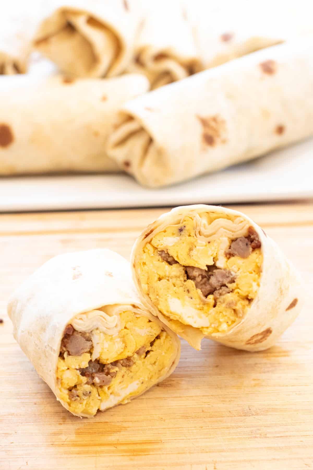 Close-up of an unwrapped campfire breakfast burrito held in two hands, showing layers of creamy scrambled eggs, crumbled sausage, melted cheddar cheese, and a drizzle of hot sauce against a blurred campfire background