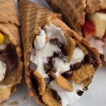 Campfire cone s'mores wrapped in foil resting over glowing campfire coals, one unwrapped to reveal melted chocolate, golden marshmallows, and fresh strawberries spilling from a crispy waffle cone