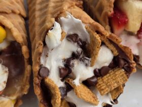Campfire cone s'mores wrapped in foil resting over glowing campfire coals, one unwrapped to reveal melted chocolate, golden marshmallows, and fresh strawberries spilling from a crispy waffle cone