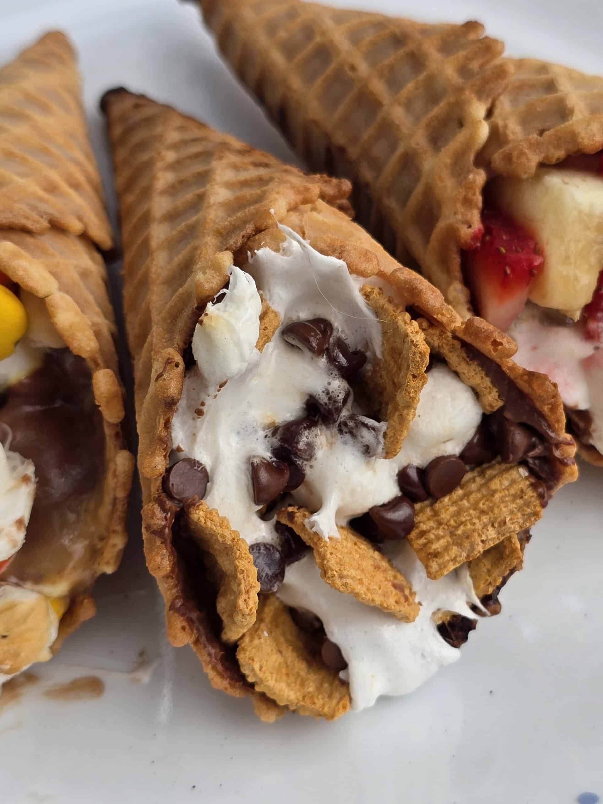 Campfire cone s'mores wrapped in foil resting over glowing campfire coals, one unwrapped to reveal melted chocolate, golden marshmallows, and fresh strawberries spilling from a crispy waffle cone