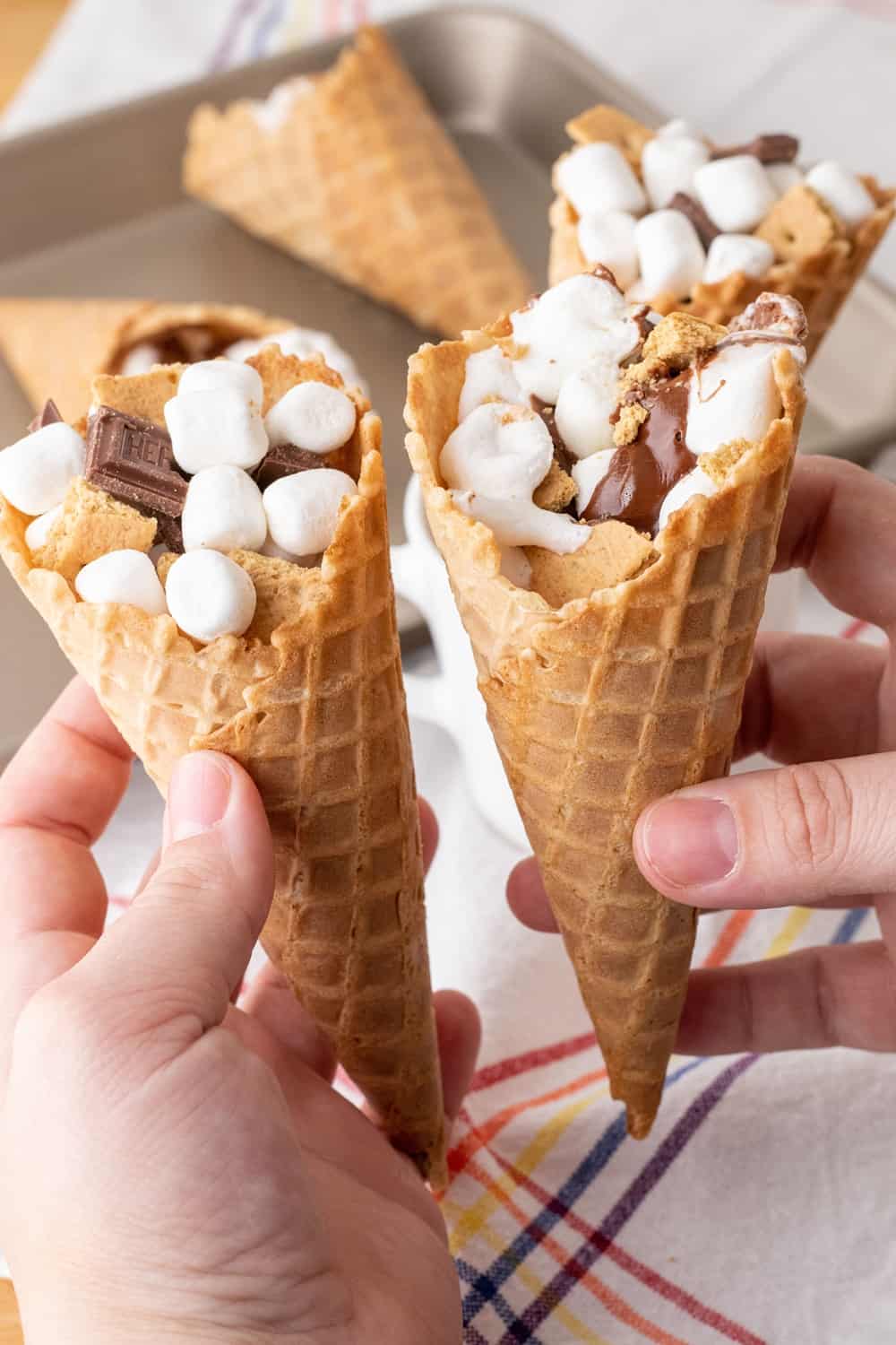 Flat lay of campfire cone s'mores ingredients — waffle cones, mini marshmallows, chocolate chips, sliced strawberries, banana slices, and squares of dark chocolate arranged on a rustic wooden board outdoors