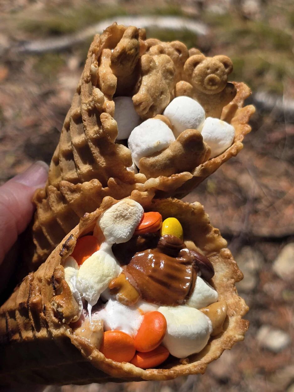 Close-up of an unwrapped campfire cone s'more held in both hands over a glowing fire, melted dark chocolate and toasted marshmallows visibly oozing from the top of a crispy waffle cone with strawberry slices