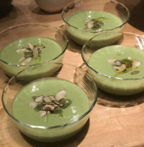 A beautifully plated bowl of chilled gazpacho with a deep red color, drizzled with olive oil, garnished with diced cucumber, cherry tomato halves, and fresh basil, shot overhead on a white linen surface with a rustic wooden spoon