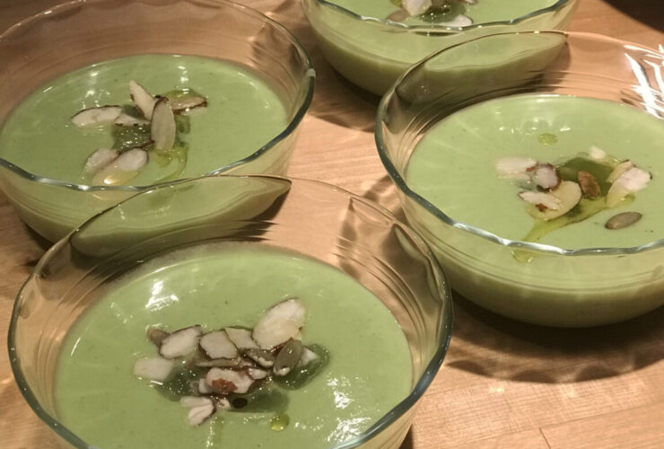 A beautifully plated bowl of chilled gazpacho with a deep red color, drizzled with olive oil, garnished with diced cucumber, cherry tomato halves, and fresh basil, shot overhead on a white linen surface with a rustic wooden spoon