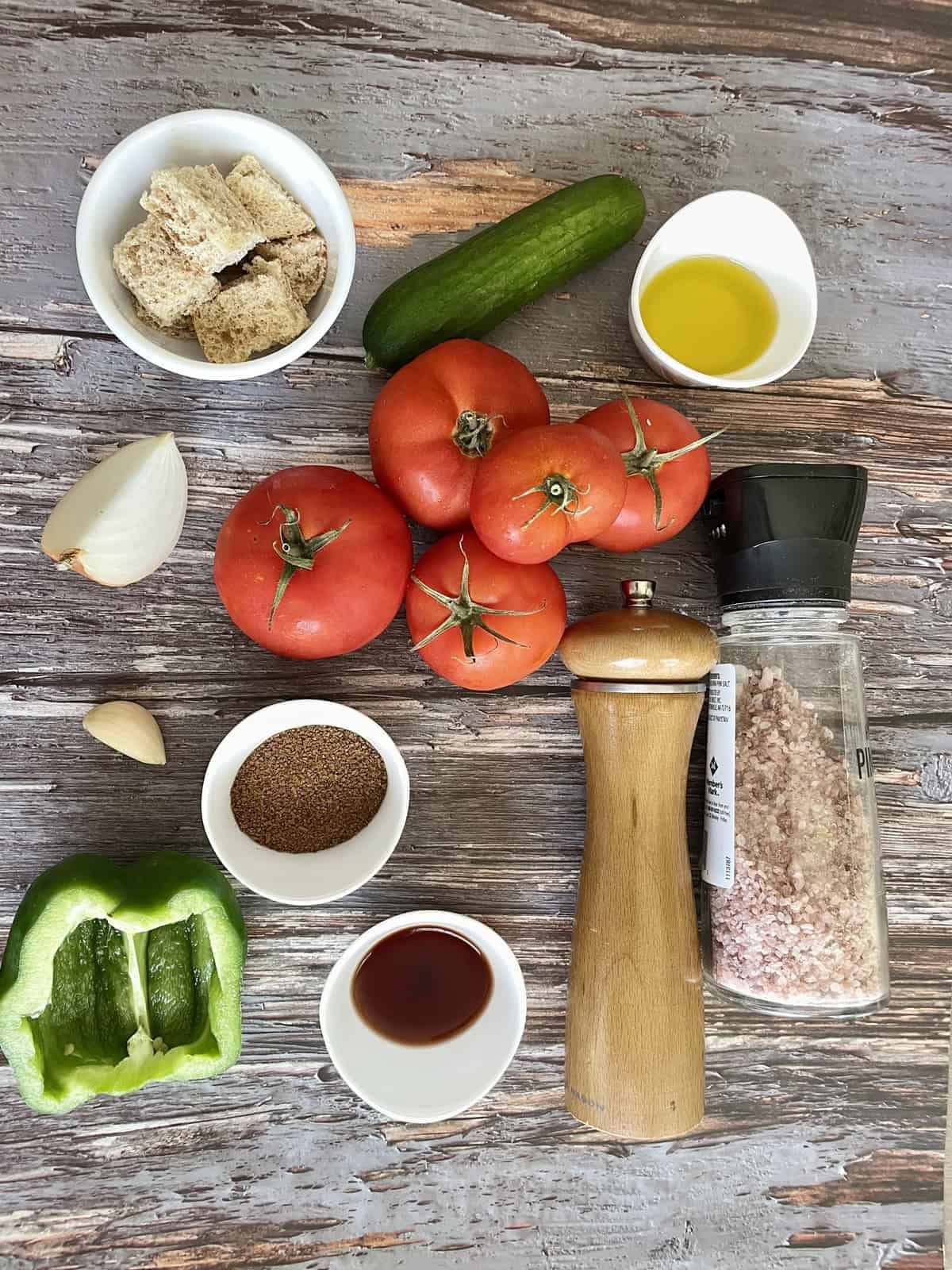 Overhead flat lay of chilled gazpacho ingredients on a marble surface — ripe red tomatoes, English cucumber, red bell pepper, garlic cloves, sherry vinegar bottle, olive oil, and fresh basil leaves arranged artfully