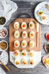 Classic deviled eggs arranged on a white marble platter, creamy yellow filling piped in neat swirls, dusted generously with smoked paprika and garnished with fresh chives, bright natural lighting, appetizing overhead shot