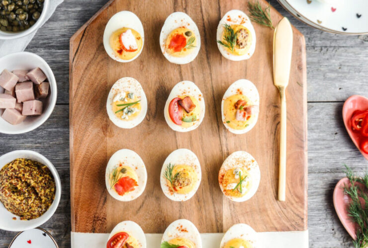Classic deviled eggs arranged on a white marble platter, creamy yellow filling piped in neat swirls, dusted generously with smoked paprika and garnished with fresh chives, bright natural lighting, appetizing overhead shot