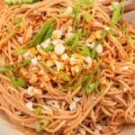 Cold sesame noodles served in a white bowl topped with sliced cucumber, shredded carrots, sesame seeds, sliced scallions, and a drizzle of chili oil, shot overhead on a dark marble surface
