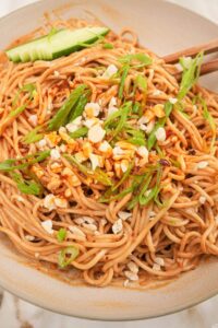 Cold sesame noodles served in a white bowl topped with sliced cucumber, shredded carrots, sesame seeds, sliced scallions, and a drizzle of chili oil, shot overhead on a dark marble surface