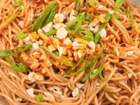 Cold sesame noodles served in a white bowl topped with sliced cucumber, shredded carrots, sesame seeds, sliced scallions, and a drizzle of chili oil, shot overhead on a dark marble surface