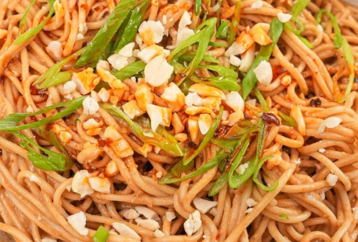 Cold sesame noodles served in a white bowl topped with sliced cucumber, shredded carrots, sesame seeds, sliced scallions, and a drizzle of chili oil, shot overhead on a dark marble surface