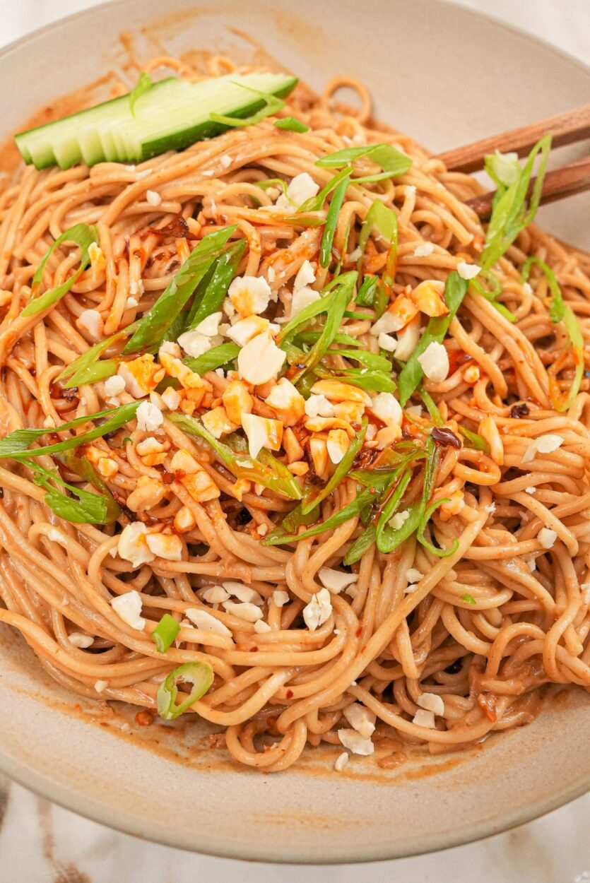 Cold sesame noodles served in a white bowl topped with sliced cucumber, shredded carrots, sesame seeds, sliced scallions, and a drizzle of chili oil, shot overhead on a dark marble surface