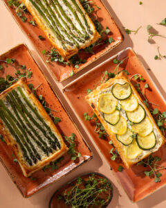 Grilled vegetable and goat cheese tart sliced on a rustic wooden board, showing charred zucchini, bell peppers, and eggplant on a creamy goat cheese filling in a golden whole wheat crust, garnished with fresh thyme and a drizzle of olive oil