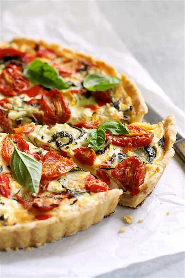 Close-up of a single slice of grilled vegetable goat cheese tart on a white ceramic plate, showing layers of charred vegetables, creamy goat cheese, and flaky whole wheat crust with fresh thyme sprigs and a drizzle of golden olive oil