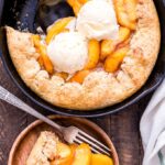 Golden peach cobbler in a rustic cast iron skillet with a flaky biscuit topping, bubbling peach filling, and a scoop of vanilla ice cream melting over the top, shot on a wooden farmhouse table