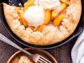 Golden peach cobbler in a rustic cast iron skillet with a flaky biscuit topping, bubbling peach filling, and a scoop of vanilla ice cream melting over the top, shot on a wooden farmhouse table