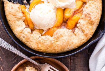Golden peach cobbler in a rustic cast iron skillet with a flaky biscuit topping, bubbling peach filling, and a scoop of vanilla ice cream melting over the top, shot on a wooden farmhouse table