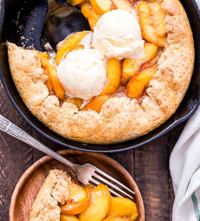 Golden peach cobbler in a rustic cast iron skillet with a flaky biscuit topping, bubbling peach filling, and a scoop of vanilla ice cream melting over the top, shot on a wooden farmhouse table