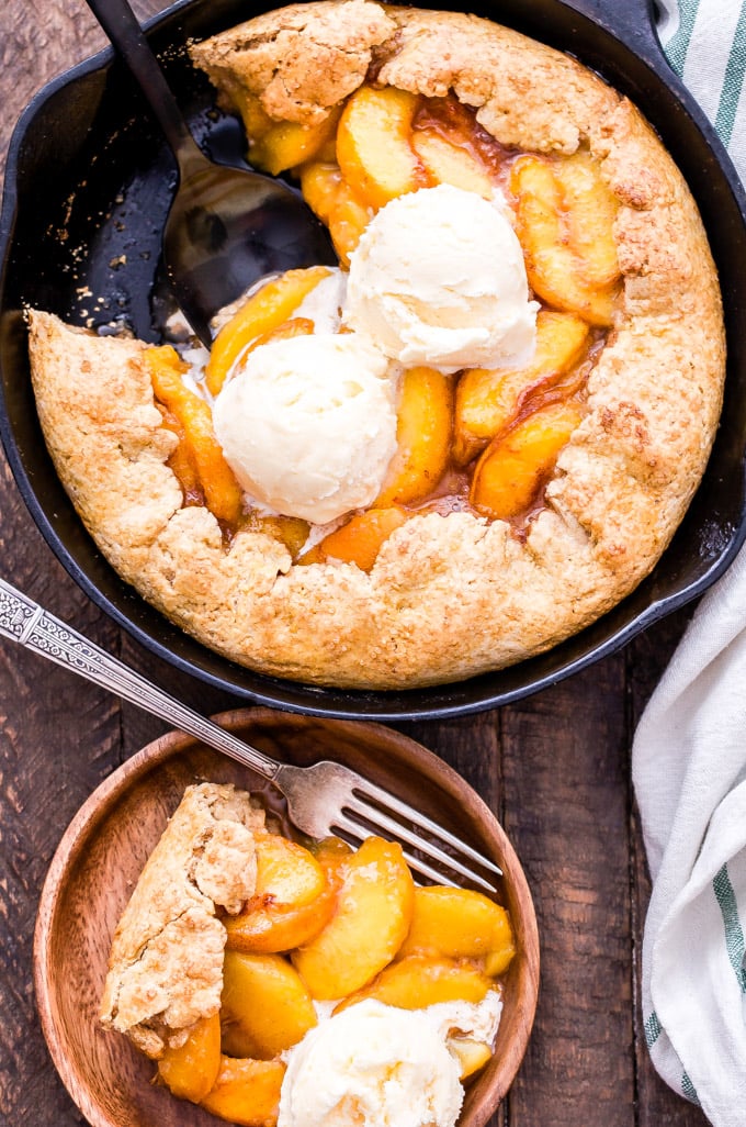 Golden peach cobbler in a rustic cast iron skillet with a flaky biscuit topping, bubbling peach filling, and a scoop of vanilla ice cream melting over the top, shot on a wooden farmhouse table
