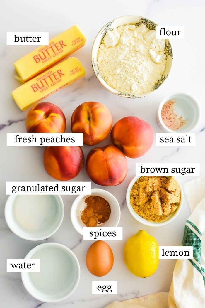 Overhead flat-lay of peach cobbler ingredients including sliced fresh peaches, cold butter cubes, brown sugar, cinnamon, flour, and heavy cream arranged on a marble surface