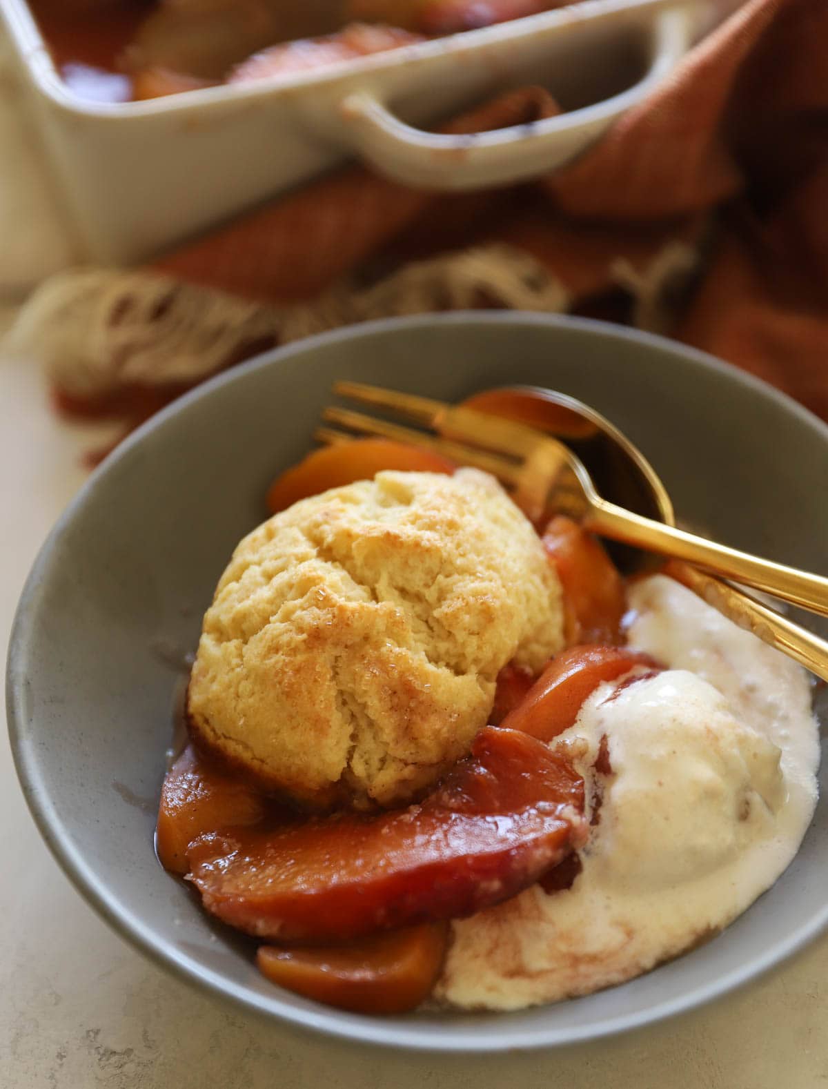 Close-up of a spoonful of warm peach cobbler being lifted from a cast iron skillet, showing the glossy caramelized peach filling and flaky golden biscuit topping with vanilla ice cream melting alongside