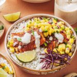 Peach mango salsa in a white bowl garnished with fresh cilantro and lime wedges, served alongside grilled fish tacos on a rustic wooden board