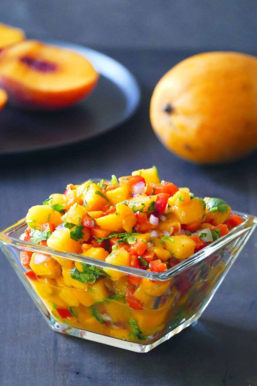 Overhead flat-lay of diced peaches, mango cubes, sliced jalapeño, chopped red onion, cilantro, and lime halves arranged on a white marble surface before mixing