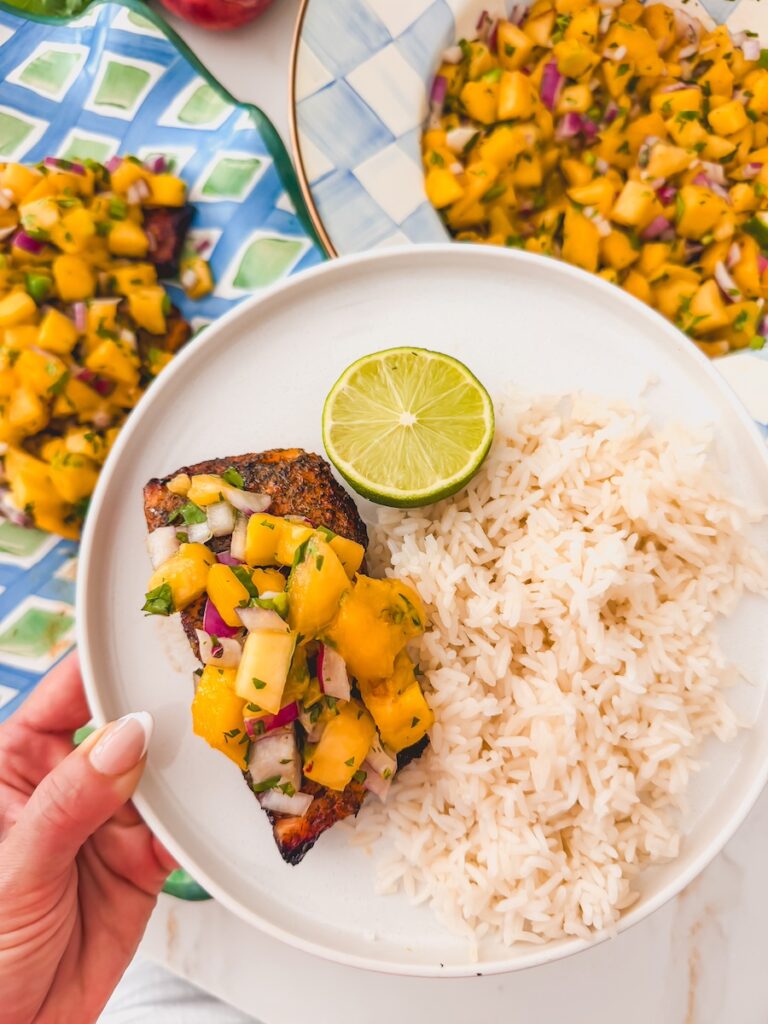 Close-up of peach mango salsa spooned generously over a seared salmon fillet on a white plate, with visible chunks of golden mango, peach, red onion, and fresh cilantro leaves
