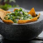 A rustic stone bowl filled with chunky restaurant style guacamole garnished with fresh cilantro leaves, sliced jalapeño, and a wedge of lime, surrounded by tortilla chips on a dark wood surface