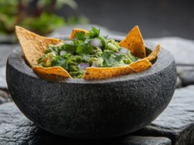 A rustic stone bowl filled with chunky restaurant style guacamole garnished with fresh cilantro leaves, sliced jalapeño, and a wedge of lime, surrounded by tortilla chips on a dark wood surface