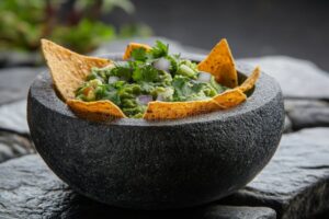 A rustic stone bowl filled with chunky restaurant style guacamole garnished with fresh cilantro leaves, sliced jalapeño, and a wedge of lime, surrounded by tortilla chips on a dark wood surface